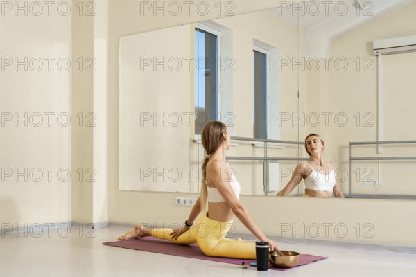 A woman practices yoga in a well-lit studio. She sits in a split position on a yoga mat while reflecting in the mirror. A singing bowl and water bottle rest nearby, enhancing her calming routine