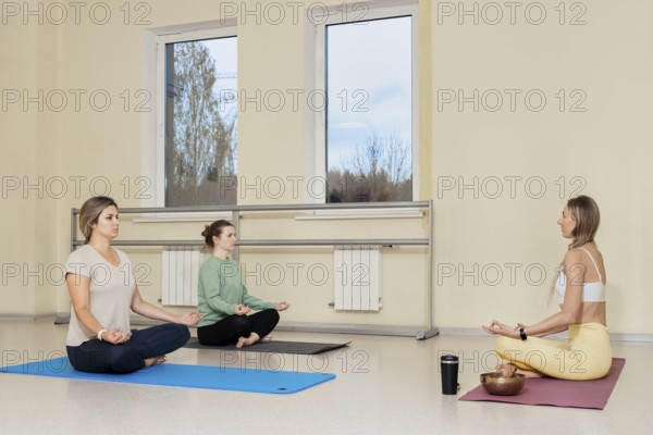 Three women are seated on yoga mats in a bright studio. They are focusing on meditation, with one instructor guiding them. The serene atmosphere promotes mindfulness and relaxation