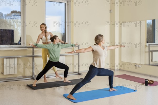 Three individuals practice yoga in a well-lit studio. One person demonstrates a warrior pose, while the others follow suit. The atmosphere is calm, promoting focus and relaxation
