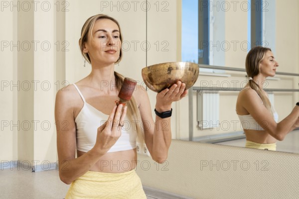 A woman stands in an airy studio, holding a singing bowl in one hand and a mallet in the other. She focuses intently, creating calming sounds as natural light enters through large windows