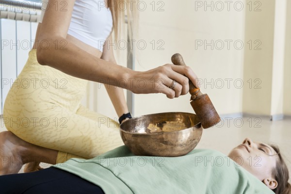 In a calm wellness studio, a sound healer plays a singing bowl on a person lying calmly on a mat. The serene atmosphere enhances relaxation and promotes healing through sound