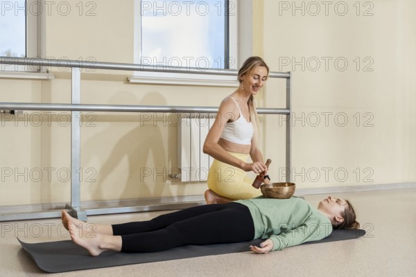 An instructor uses a singing bowl to provide sound therapy to a relaxed participant lying on a mat in a bright studio. The peaceful setting enhances the calming experience for both