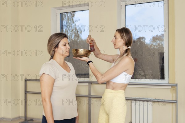 In a serene studio, a woman stands calmly while another person gently uses a singing bowl above her head. The soothing sound promotes relaxation and mindfulness in a peaceful environment