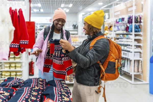 Happy young multiracial couple smiling as they browse and select red holiday sweaters in a busy retail store, enjoying festive shopping, bonding and seasonal gift buying