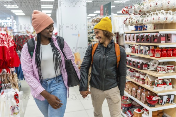 Diverse young couple smiling while holding hands, shopping together in a store, browsing shelves filled with christmas and holiday decorations and gifts