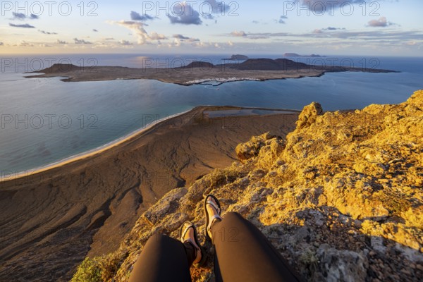 First person view, legs of a woman sitting on a steep cliff, view of the sea and the island of La Graciosa, Mirador del Porrito viewpoint at sunset, Lanzarote, Canary Islands, Spain