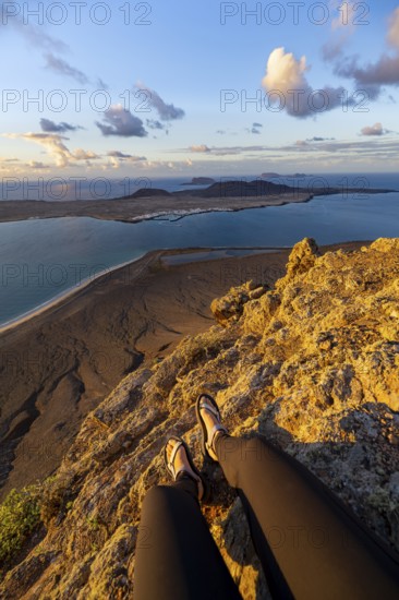 First person view, legs of a woman sitting on a steep cliff, view of the sea and the island of La Graciosa, Mirador del Porrito viewpoint at sunset, Lanzarote, Canary Islands, Spain