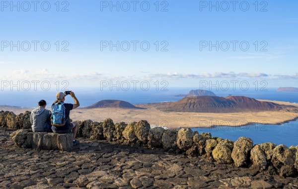 Tourists on a bench on the viewing platform at the Mirador del RÃ­o viewpoint, in the evening light, view of the sea and of the island of La Graciosa with volcanic craters, designed by artist César Manrique, Lanzarote, Canary Islands, Spain