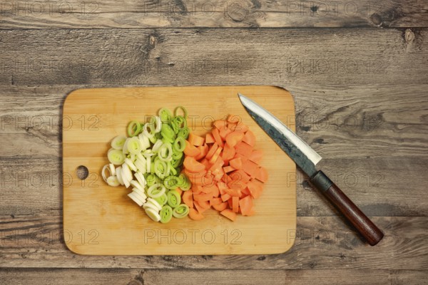 Fresh leek onion and carrots are chopped and neatly arranged on a wooden cutting board. A sharp knife rests beside, indicating readiness for cooking and meal preparation