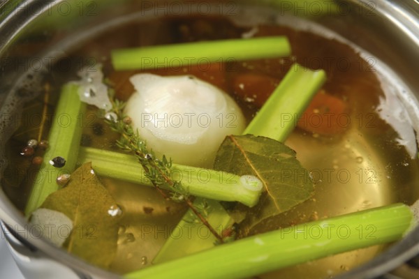 A pot simmers on the stove filled with water, celery stalks, onions, and spices. Fresh herbs like thyme and bay leaves add aroma, creating a hearty broth. The scene captures simple cooking at home