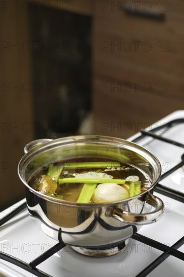 Cooking vegetable broth on the stove in a stainless steel pot with fresh celery, onion, carrot, bay leaf and peppercorns. Steam rises from the pot, indicating a warm cooking environment during meal preparation