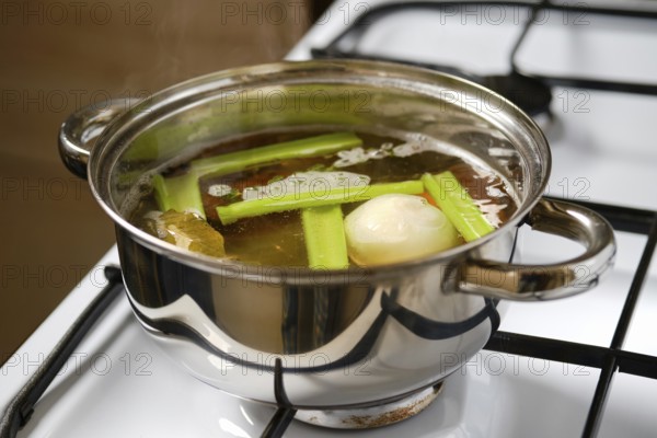 Fresh celery, carrot and an onion simmer in a pot of broth on the stovetop. The pot shows steam rising as the ingredients cook, creating a warm, inviting scene in a kitchen setting