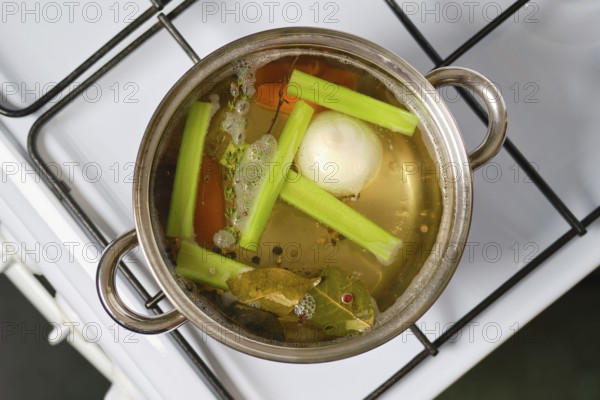 A silver pot sits on a stove filled with bubbling water, celery stalks, and seasonings. Ingredients like onion and bay leaves are present, creating a flavorful vegetable broth as it cooks