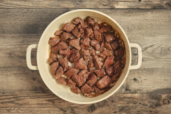 Chunks of beef are searing in a large pot on a weathered wooden table. The meat is cooked to a rich brown color, suggesting a flavorful dish is being prepared