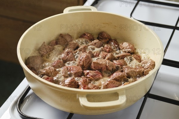 Pieces of beef are sizzling and cooking in a large pot on a gas stove. The meat is browning while juices bubble, showcasing a delicious preparation process for a hearty meal
