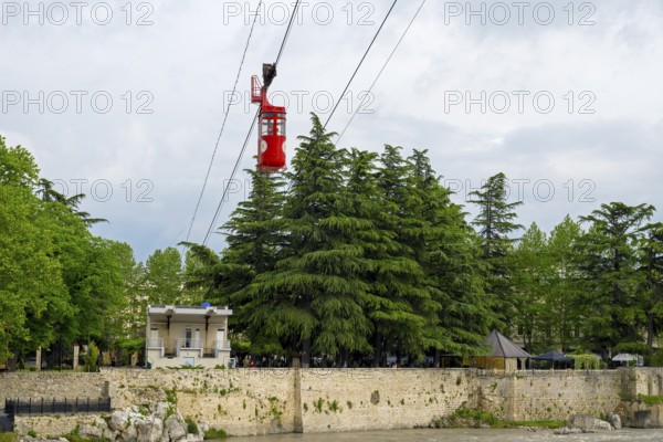 A red cable car cabin above tree tops near a river under a cloudy sky, cable car station, Kutaisi, Imereti region, Georgia