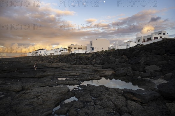 Black coast of volcanic rocks behind typical white houses of the village of La Santa, at sunset, Lanzarote, Canary Islands, Spain