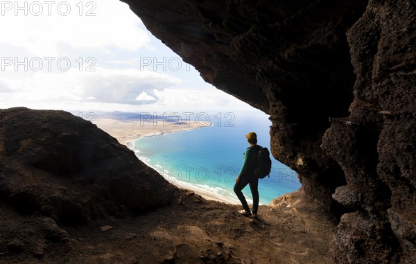 Tourist in the Cueva de las Cabras cave, young woman enjoying the view from the Risco de Famara cliffs on Famara beach, Playa de Famara with La Calaeta, Lanzarote, Canary Islands, Spain