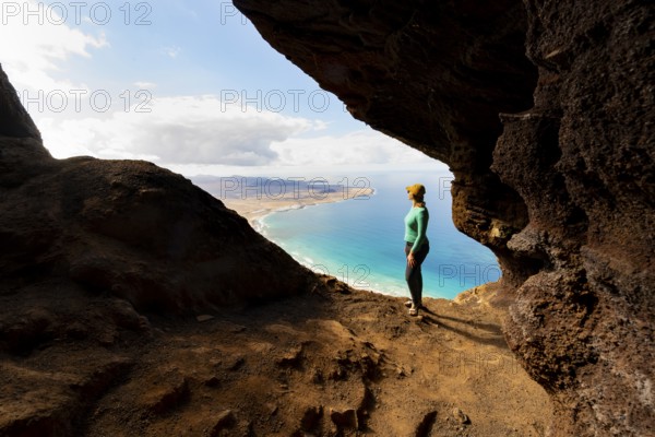 Tourist in the Cueva de las Cabras cave, young woman enjoying the view from the Risco de Famara cliff to Famara beach, Playa de Famara with La Calaeta, Lanzarote, Canary Islands, Spain