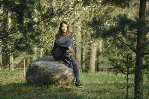 A young woman is seated on a large rock in the midst of a vibrant green forest filled with blooming trees. The sunlight filters through the trees, creating a serene and peaceful atmosphere