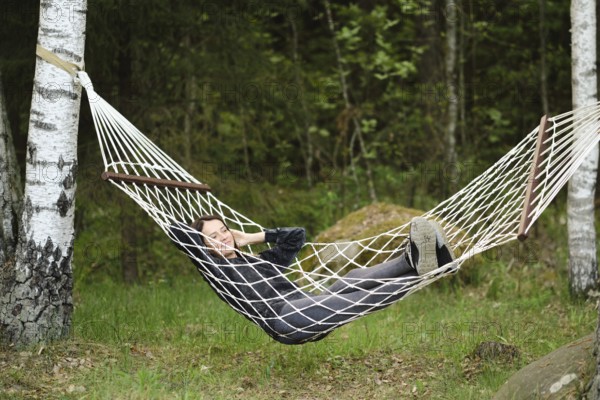 A woman enjoys a peaceful afternoon in a hammock, nestled between trees. She has a relaxed posture, with the serene green landscape creating a calming atmosphere in the woods