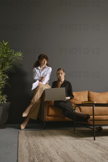 Two women focus on their work as they share ideas and discuss projects while seated in an office sofa. One woman stands beside the couch, and the other sits with a laptop, surrounded by greenery