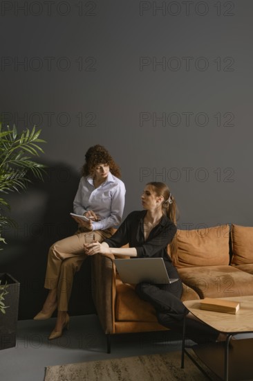 In a stylish living room, two women engage in a collaborative work meeting. One writes notes while the other discusses ideas, seated on a cozy couch