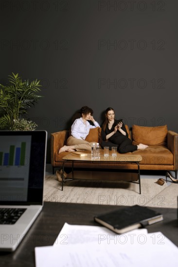 Two individuals are seated comfortably on a stylish couch, one looking at a phone while the other gazes thoughtfully. They are situated in a contemporary living room with a dark wall