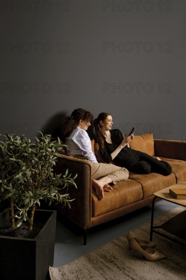 Two friends sit comfortably on a brown couch in a modern living room, engaged with a mobile device. They enjoy each other's company, surrounded by minimalist decor and indoor plants