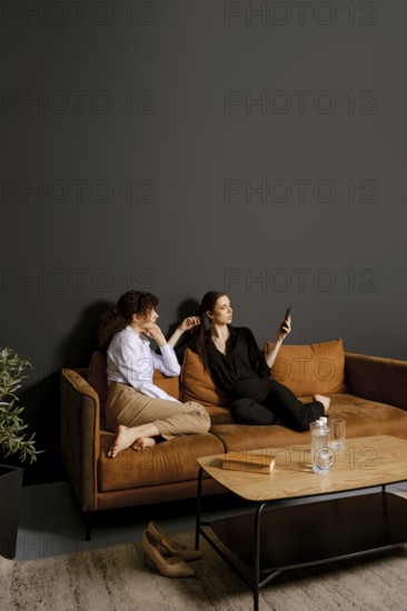 Two friends sit on a comfortable couch in a stylish living room. One is scrolling on a smartphone while the other watches with a relaxed expression, creating a warm and inviting atmosphere