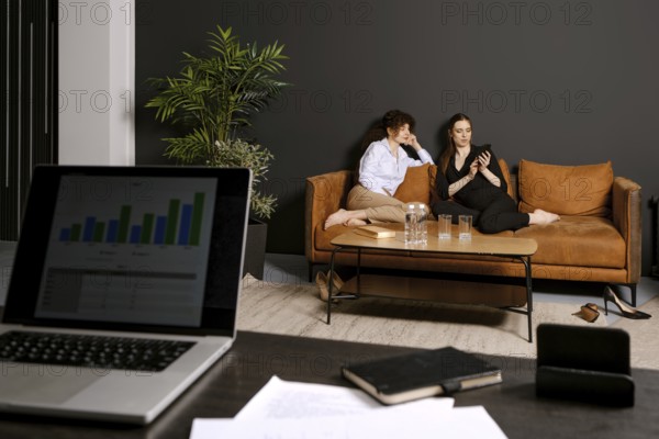 Two friends sit comfortably on a brown couch in a stylish living room, engaging with a smartphone. A laptop and documents are visible on a nearby table, suggesting a casual work environment