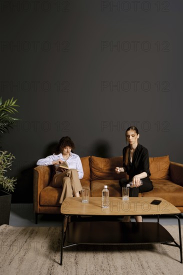 In a stylish indoor setting, two individuals sit on a brown couch. One person reads a book while the other holds a cup, engaged in conversation. A plant and water glasses complete the scene