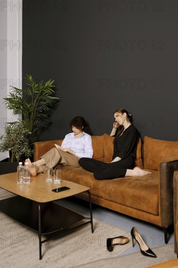 Two friends enjoy a cozy afternoon on a comfortable sofa in a modern living room. One reads intently while the other chats on the phone, creating a warm and casual atmosphere