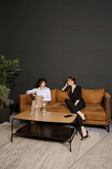 Two individuals engage in a conversation while sitting in a contemporary living room. The setting features a brown sofa and a coffee table