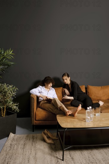 Two friends sit comfortably on a warm brown couch in a stylish room, engaging in conversation. Lush greenery and a sleek coffee table with water bottles enhance the inviting atmosphere