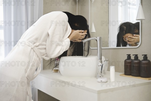 A woman in a white bathrobe leans over a stylish sink in a contemporary bathroom, splashing water on her face