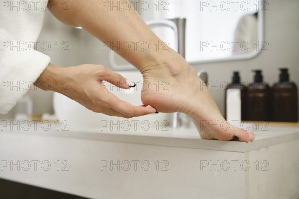 A person gently massages their foot while resting on a sink in a stylish bathroom. Soft morning light enhances the serene atmosphere. Dark bottles are neatly arranged on the counter nearby