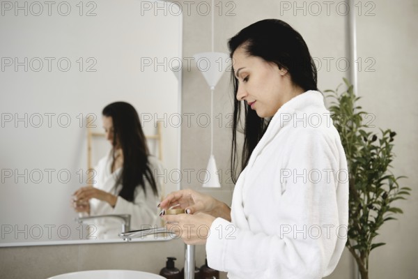 A woman with long dark hair stands at a sleek sink in a contemporary bathroom and opens a jar of cream