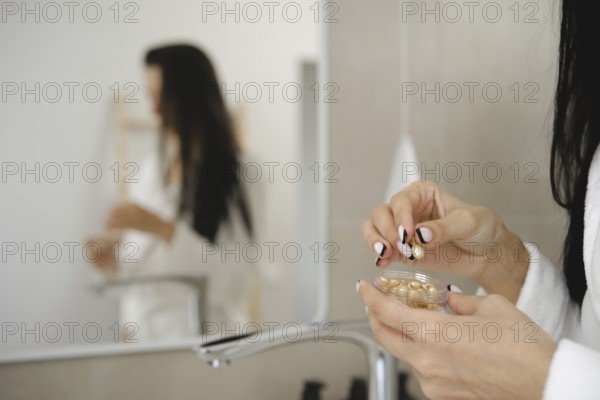 In a stylish bathroom, a woman in a robe is holding a small jar with skin vitamins and preparing to apply them