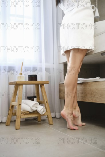 Unrecognizable woman in white robe standing barefoot on tiptoe in a well-lit bathroom