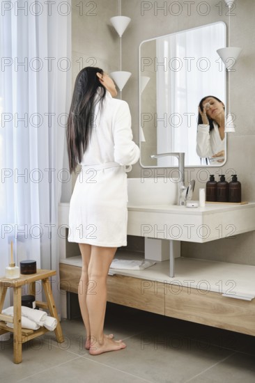 Barefoot woman wearing a white robe stands by a bathroom sink, facing the mirror. She appears to be preparing for her day, surrounded by a clean and contemporary setting with soft lighting
