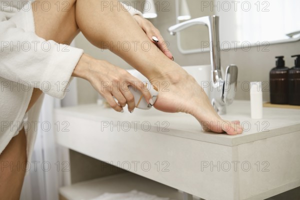 A woman in a bathrobe delicately clean her foot at a sleek bathroom sink with electric exfoliator