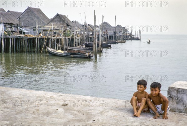 Two boys sitting on quayside next to attap houses of fishing village on stilts, Singapore, southeast Asia 1963