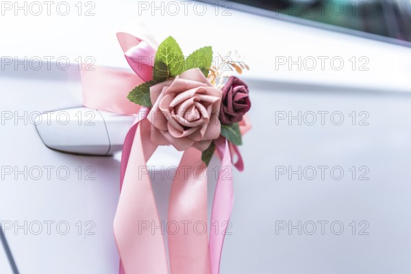 Elegant pink ribbons and artificial flowers adorn the handle of a white car, signifying a wedding celebration