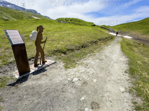 Information board on the left next to the Alpine Pass border pass road on early history and pass crossing in ancient times from the Alpine Pass border pass Little St. Bernard, next to it a stylised hiker with a walking stick and rucksack, on the right an old mule track parallel to the pass road, Aosta Valley region, Italy