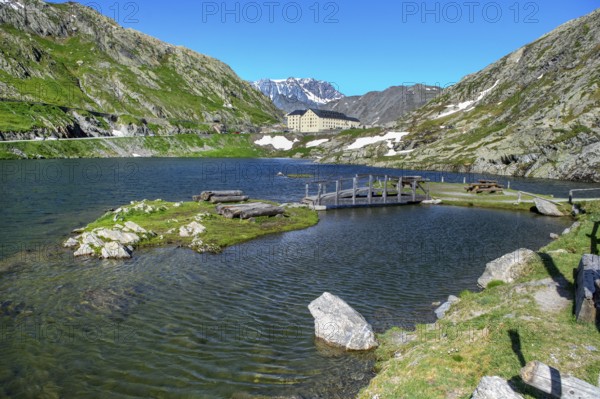 View of small island and simple wooden bridge in alpine lake Lac du Grand Gd Saint St Bernard, in the background building with hostel Auberge and hospice Hospice on alpine pass Großer Sankt St Bernhard Col du Grand Gd Saint St Bernard Colle del Gran San Bernardo, region Valle d'Aosta, Italy, canton Valais, Switzerland