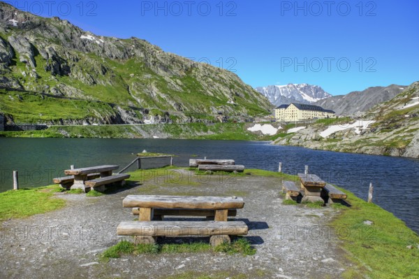 View of small island with wooden benches in mountain lake Alpine lake Lac du Grand Gd Saint St Bernard, in the background building with hostel Auberge and hospice Hospice on Alpine pass Great Saint Bernard Col du Grand Gd Saint St Bernard Colle del Gran San Bernardo, region Valle d'Aosta, Italy, canton Valais, Switzerland