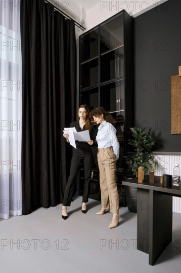 Two professional women stand in a sleek office, reviewing important documents. Sunlight filters through sheer curtains, highlighting their focused expressions as they collaborate on a project