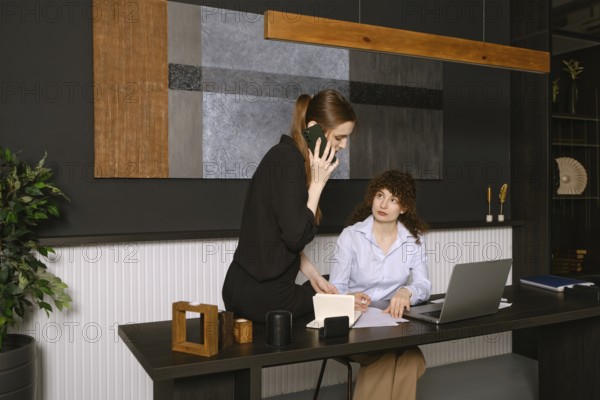 Two women are engaged in a work discussion in a stylish office setting. One is seated at a desk with a laptop, while the other stands beside her on the phone, sharing ideas and collaborating