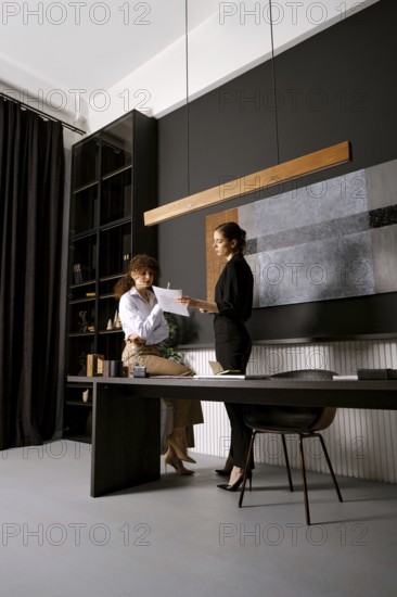 Two women are engaged in a discussion in a contemporary office space, surrounded by dark walls and elegant decor